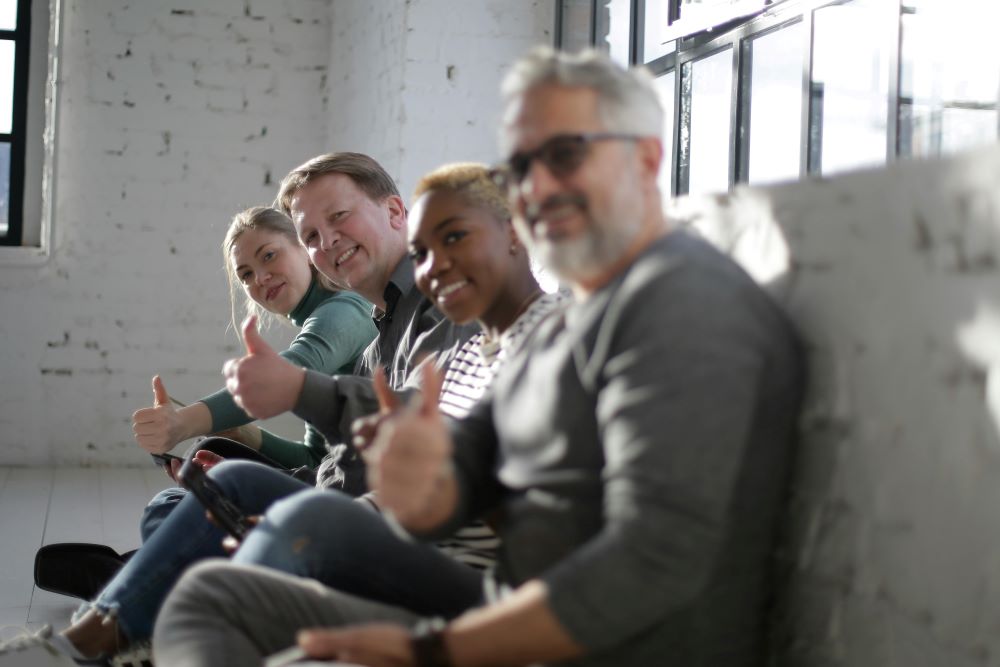 A diverse group are sat together looking towards the camera and smiling doing thumbs up.